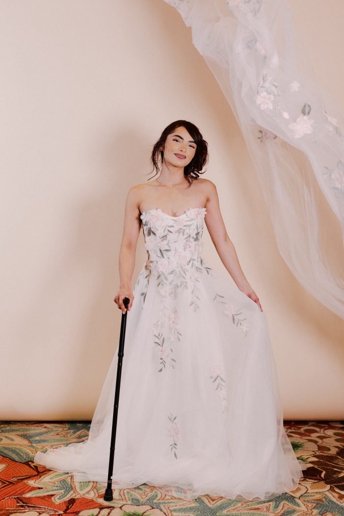 Blossom wedding dress by Rebecca Ingram featuring a strapless sweetheart neckline with delicate pastel floral appliqués across the bodice. The gown flows into a soft tulle skirt with scattered embroidered florals, styled with a sheer embroidered veil, with the model holding a cane in a neutral studio setting.
