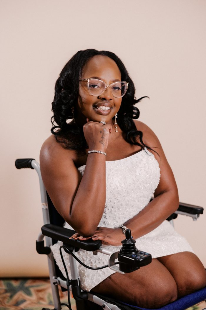 A bride wearing the Taylor gown by Rebecca Ingram smiles warmly while seated in a power wheelchair against a soft neutral background. The strapless mini-length gown features allover pearl and sequin beading. She accessorizes with clear cat-eye glasses, dangling pearl earrings, a diamond tennis bracelet, and a sparkling ring, with long black curly hair framing her face.