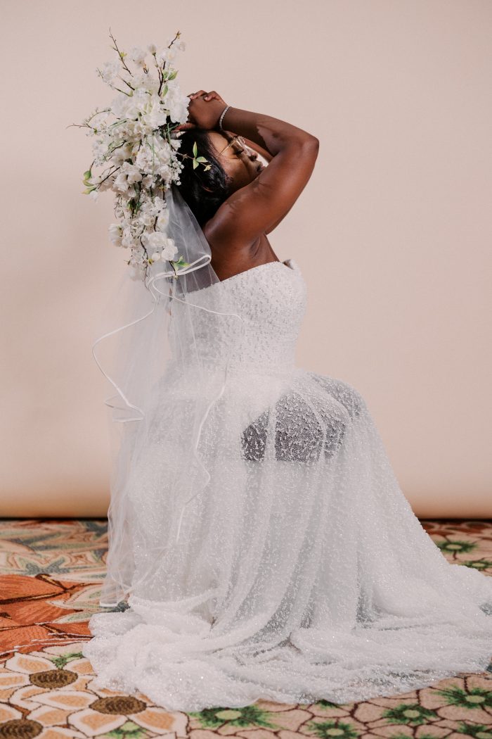 A bride wearing the Taylor gown by Rebecca Ingram poses dramatically on the floor, arms raised overhead holding a lush cascading bouquet of white orchids, roses, and flowering branches alongside a sheer veil. The strapless ballgown silhouette is covered in allover glittering sequin and beading that sparkles from the fitted bodice through the full sweeping train, pooling across a patterned carpet.