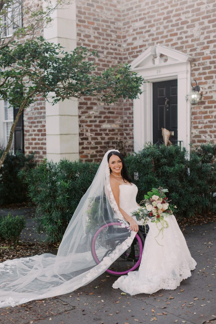 Person seated in a wheelchair outdoors in front of a brick building and greenery, wearing the Verina wedding dress by Maggie Sottero—a lace ballgown with a long sheer veil—holding a bouquet beside a wheelchair with purple wheels and an extended train cascading onto the ground.