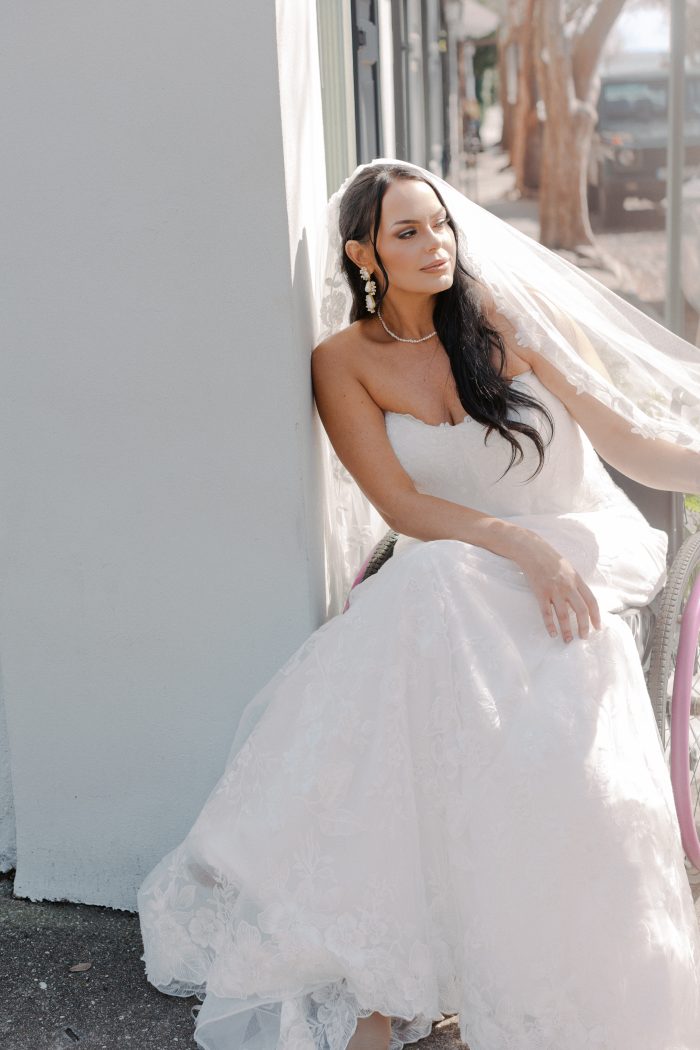 Meredith wedding dress by Maggie Sottero in a close-up view, highlighting the strapless lace bodice and softly textured skirt. The look is styled with a sheer veil as the model sits in a wheelchair, posed against a sunlit exterior wall.