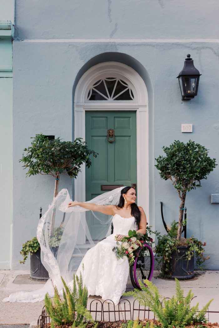 Meredith wedding dress by Maggie Sottero featuring a strapless sweetheart neckline with delicate lace appliqués over a fitted bodice. The gown flows into a soft A-line skirt, styled with a long veil and bouquet, with the model seated in a wheelchair outside a pastel blue doorway framed by greenery.