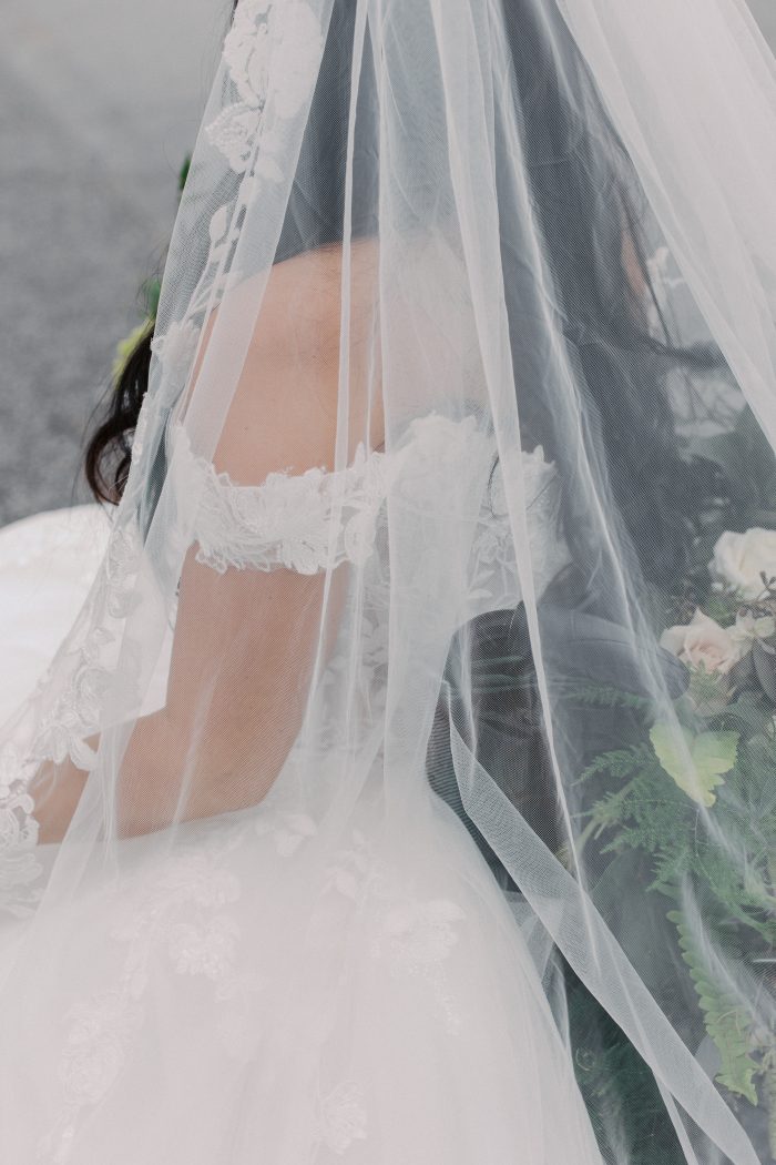 Verina wedding dress by Maggie Sottero in a close-up detail shot, featuring a sheer veil cascading over delicate lace straps and bodice with soft floral appliqués. The airy tulle layers partially obscure the gown while revealing its romantic texture and intricate detailing.