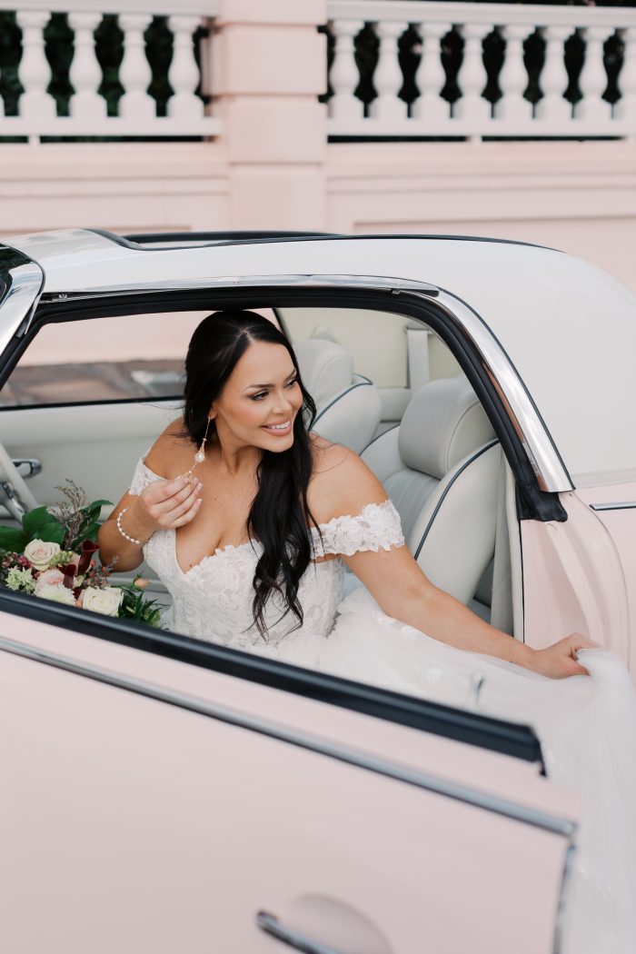 Person seated in a vintage convertible with a bouquet while wearing the Verina wedding dress by Maggie Sottero, an off-the-shoulder lace gown, with a soft pink exterior and classic white interior surrounding the scene.