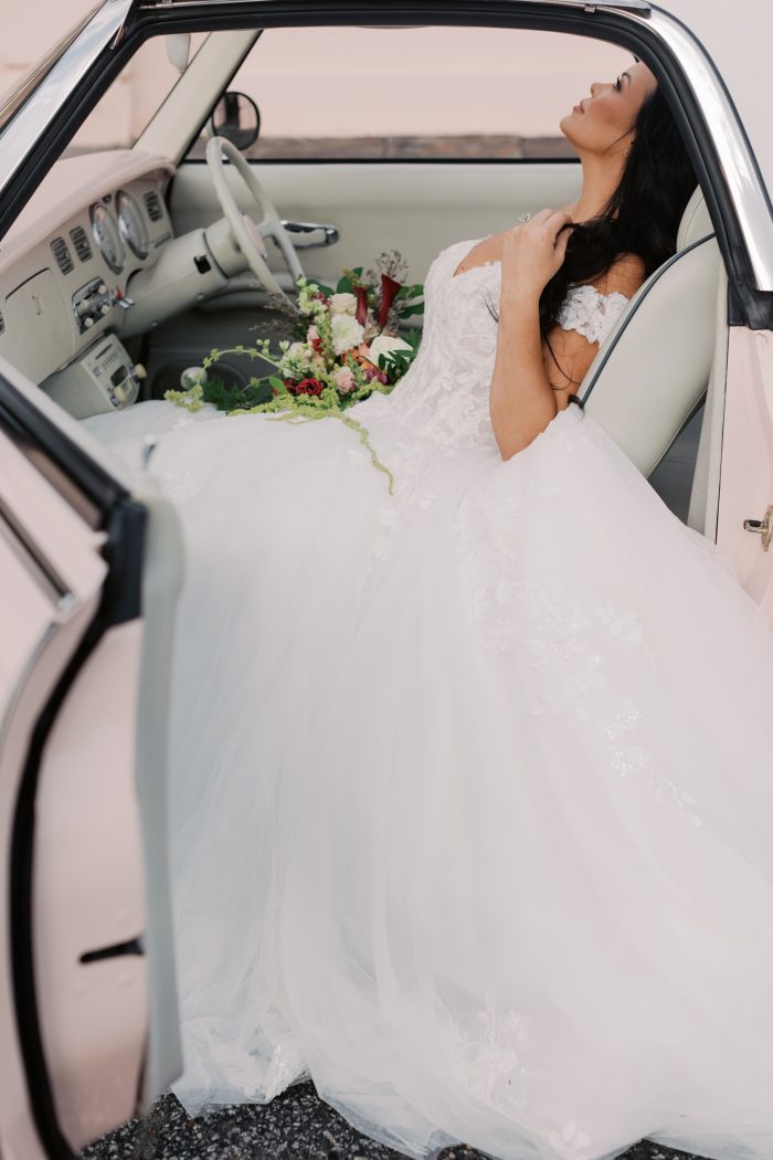 Verina wedding dress by Maggie Sottero in a lifestyle setting, featuring an off-the-shoulder lace bodice with floral appliqués and a softly structured silhouette. The look is styled with bridal accessories while seated in a vintage convertible, holding a bouquet against an elegant outdoor backdrop.