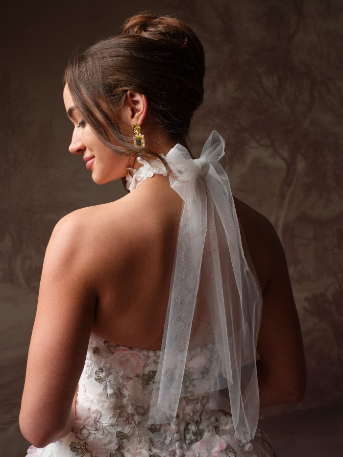 Back view detail of the Maggie Sottero Benton wedding dress choker, styled as a tulle bow tied at the nape of the neck with trailing ribbon ends. The lace and floral choker base is visible at the neck. The back of the Benton gown is shown below, featuring pink and ivory 3D floral and embroidered lace appliqués. Styled with a high bun and gold crystal drop earrings