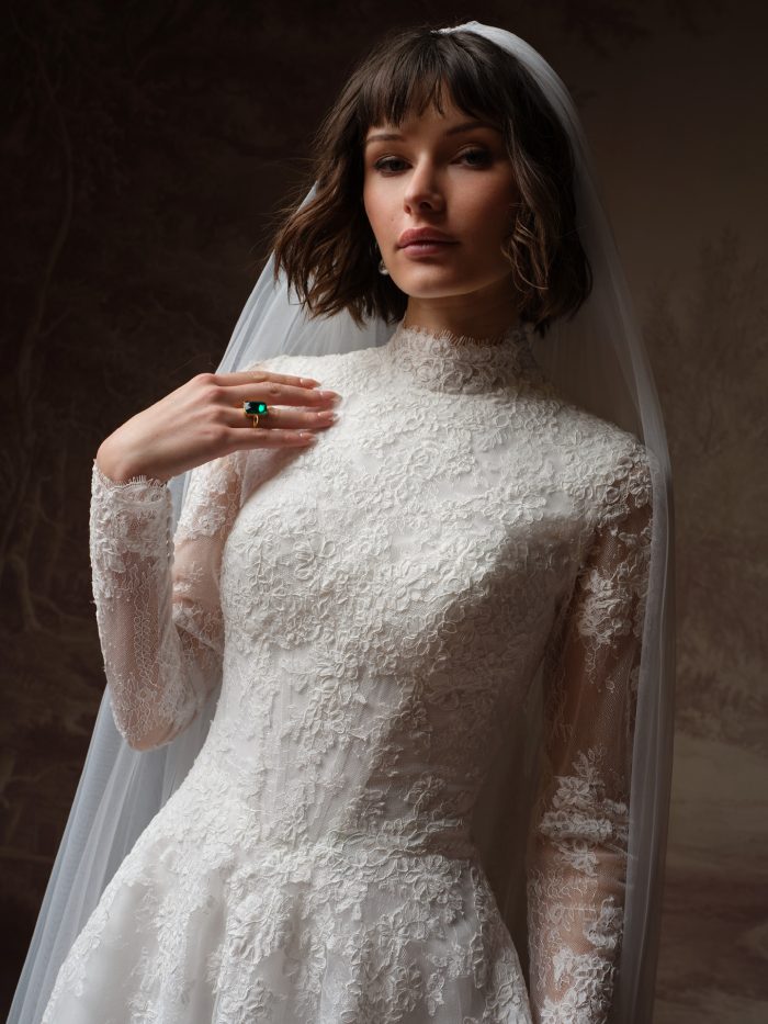 Dominique ballgown wedding dress by Maggie Sottero in a close-up view highlighting the high lace neckline, sheer long sleeves, and intricate floral lace texture across the bodice and upper skirt.