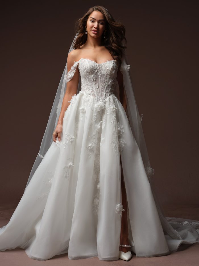 Maggie Sottero Fern A-line wedding dress in ivory, featuring a sweetheart neckline with detachable off-the-shoulder sleeves, sheer boned corset bodice, and scattered 3D floral appliqués throughout. The full tulle skirt includes a front slit and chapel train. Paired with a matching cathedral veil, pearl ankle strap heels, and gold drop earrings.