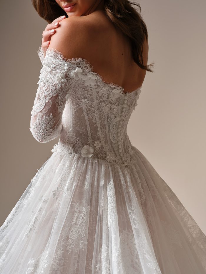Back view detail of the Maggie Sottero Kitaro, highlighting the sheer lace off-the-shoulder sleeve with eyelash trim and 3D floral appliqué cascading across the shoulder and illusion corset bodice.