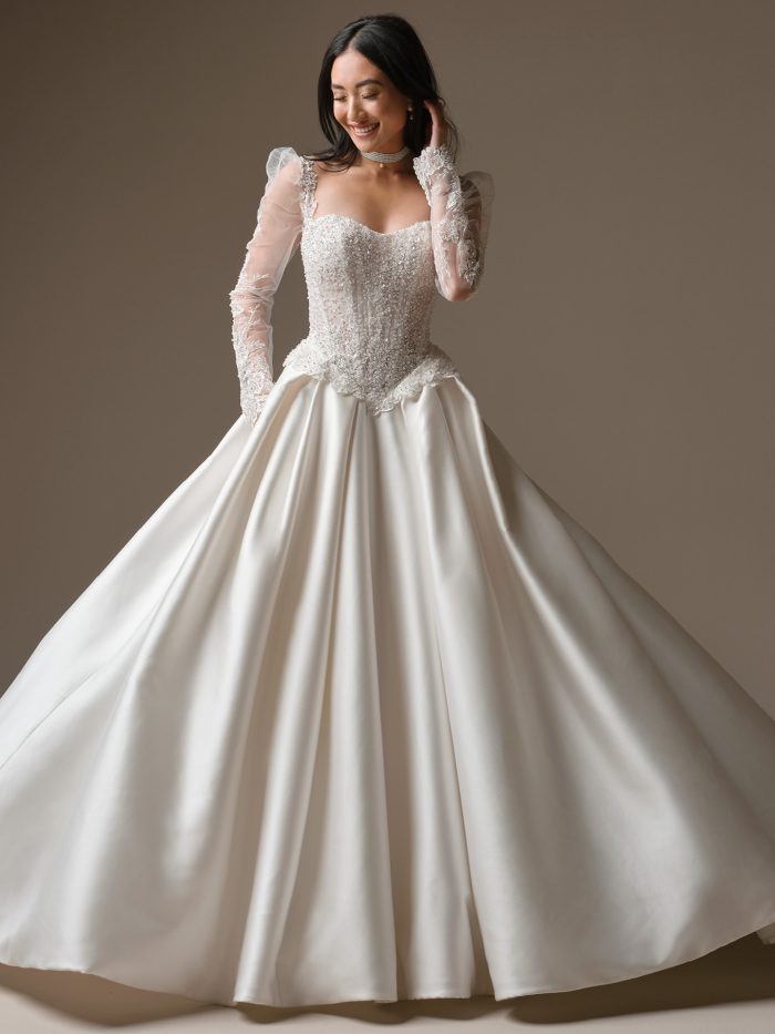 Maggie Sottero London ballgown wedding dress in ivory, featuring a sweetheart neckline, heavily beaded and lace corset bodice with a scalloped peplum waist, sheer puff-shoulder long sleeves with floral lace appliqués, and a voluminous smooth satin ballgown skirt in motion. Styled with a multi-strand pearl choker and drop earrings.
