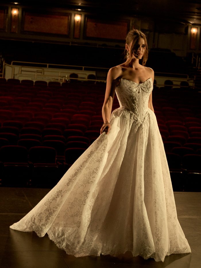Ryland ballgown wedding dress by Maggie Sottero with a strapless, beaded lace bodice and full flowing skirt with an overskirt train, shown in a full-length view on a theater stage with rows of red seats in the background.