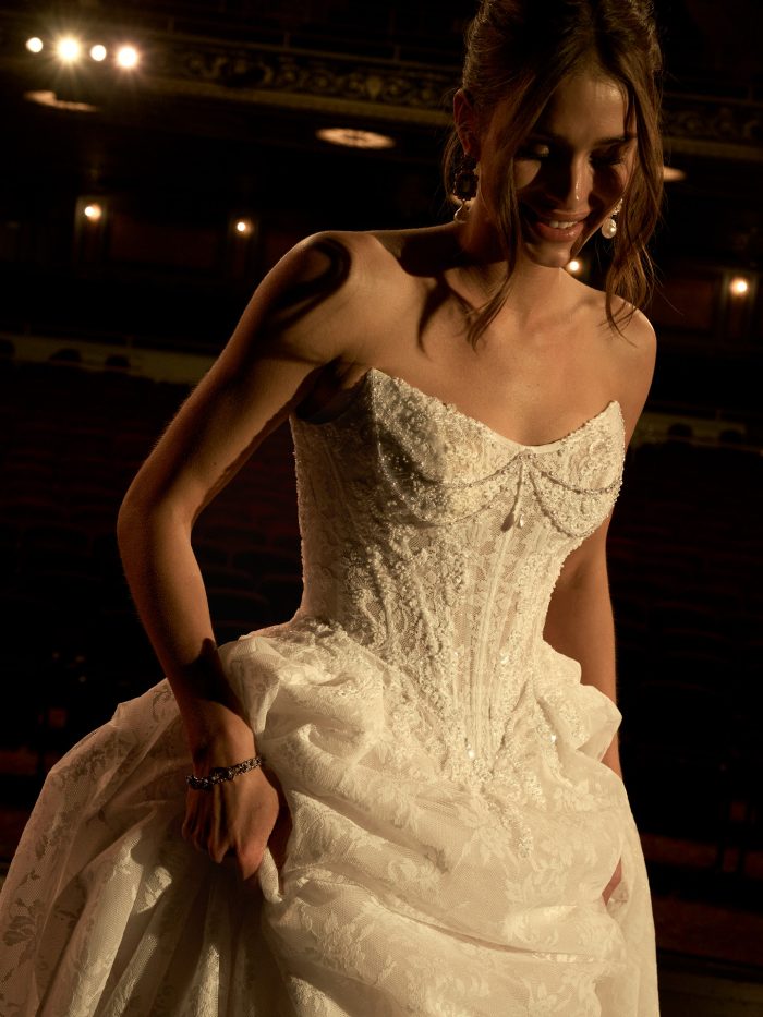 Ryland ballgown wedding dress by Maggie Sottero featuring a strapless, lace corset bodice with intricate beading and pearl draping, shown in a close-up under warm stage lighting with theater seating in the background.