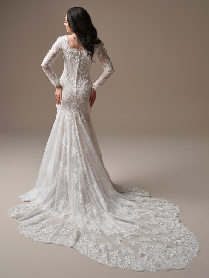 Back view of the Maggie Sottero Takera Leigh fit-and-flare wedding dress, highlighting sheer lace long sleeves with floral appliqué, a button-down back on the illusion corset bodice, and a dramatic cathedral train with intricate eyelash lace trim.