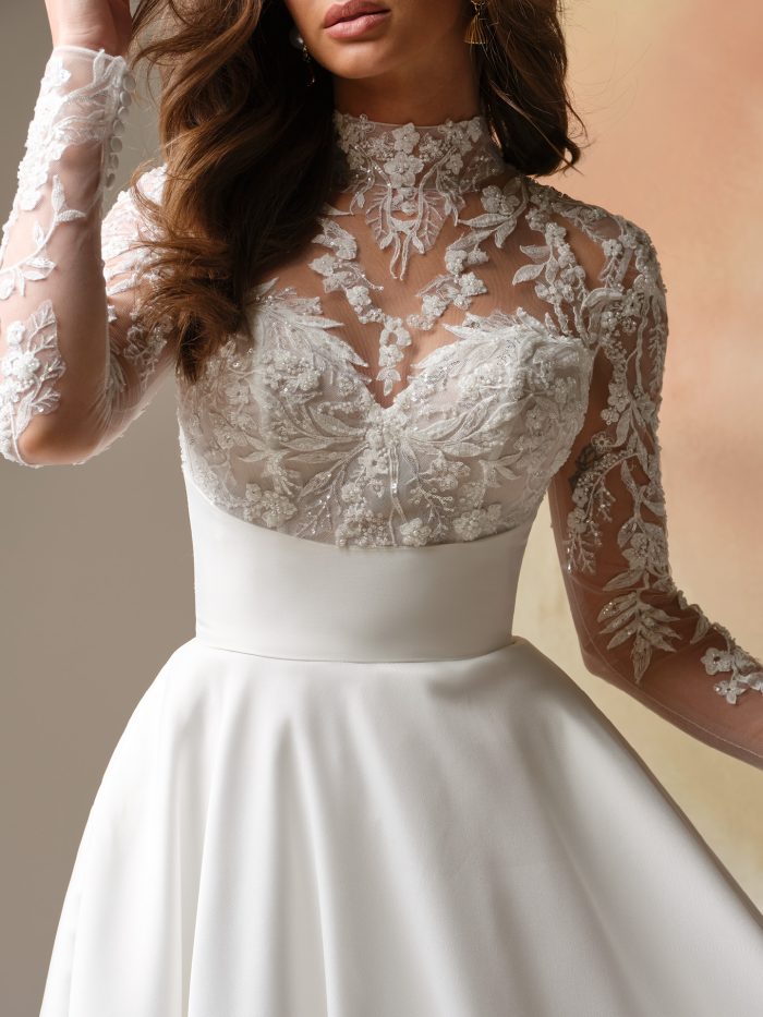 Trinity A-line wedding dress by Maggie Sottero in a close-up view highlighting the illusion lace neckline, long sheer sleeves with floral appliqués, and detailed lace bodice layered over a sweetheart lining.