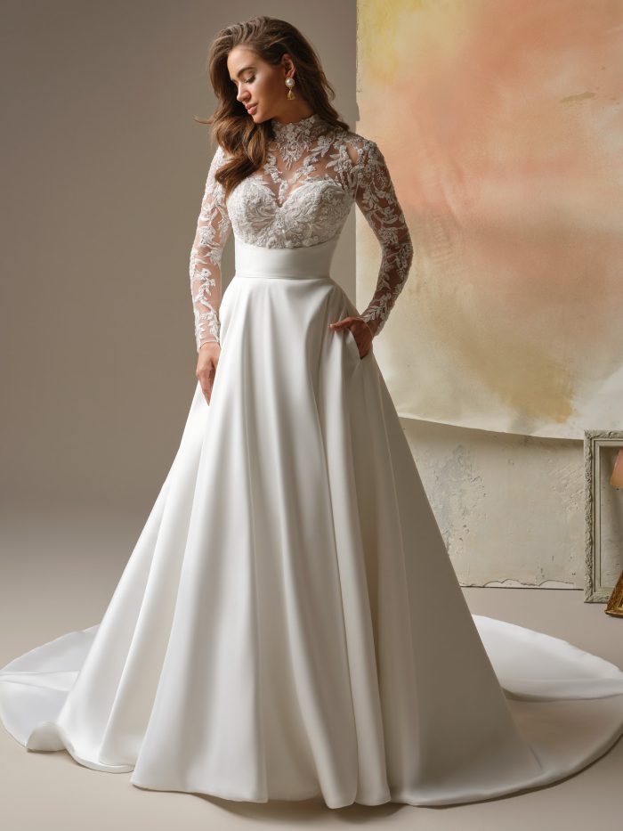Trinity A-line wedding dress by Maggie Sottero featuring an illusion high neckline with intricate floral lace over a sweetheart bodice and sheer long sleeves. The structured waist flows into a smooth A-line skirt with soft volume, styled with pockets against a warm, neutral studio backdrop.
