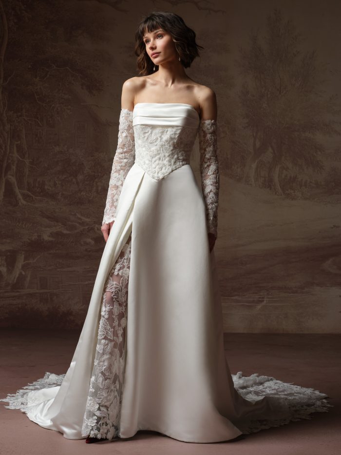 Maggie Sottero Wisteria A-line wedding dress in ivory, featuring a strapless folded satin neckline, sheer beaded and embroidered lace corset bodice, and detachable long lace sleeves. The smooth satin overskirt parts at the front to reveal a sheer floral lace underskirt, with both layers trailing into a chapel train with lace hem. Photographed against a painted mural backdrop.
