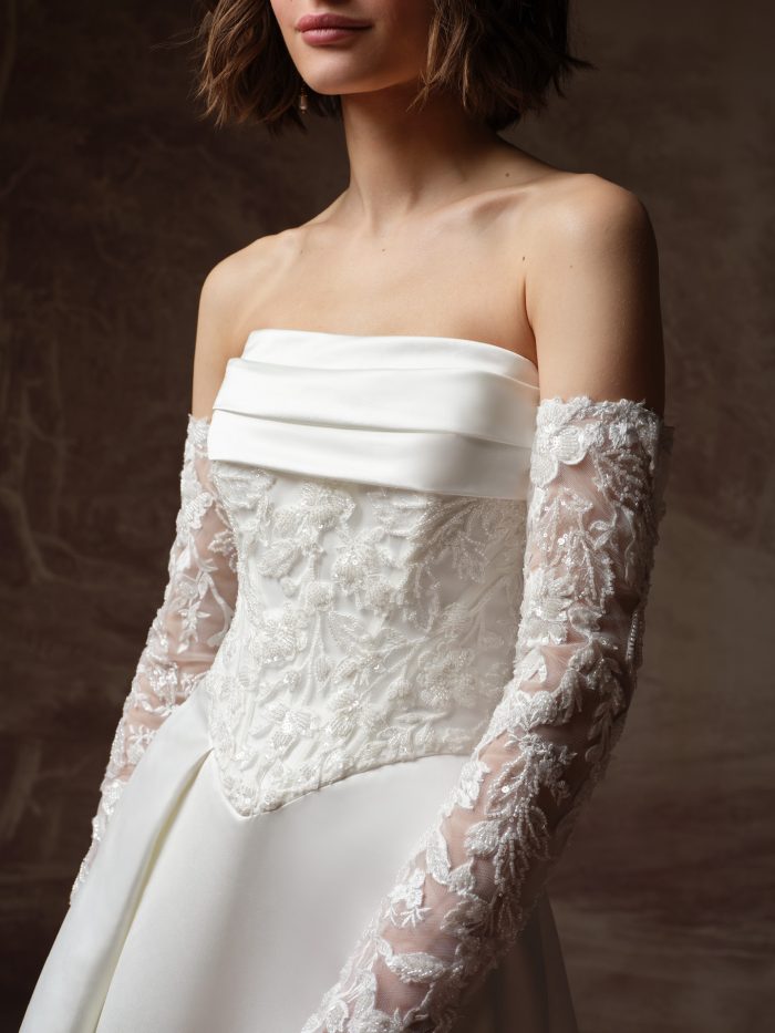 Close-up detail of the Maggie Sottero Wisteria wedding dress, highlighting the folded satin strapless neckline, sheer beaded floral and leaf lace corset bodice, and the detachable long lace sleeves featuring sequined floral and vine embroidery on a sheer tulle base. The smooth satin overskirt is visible below.