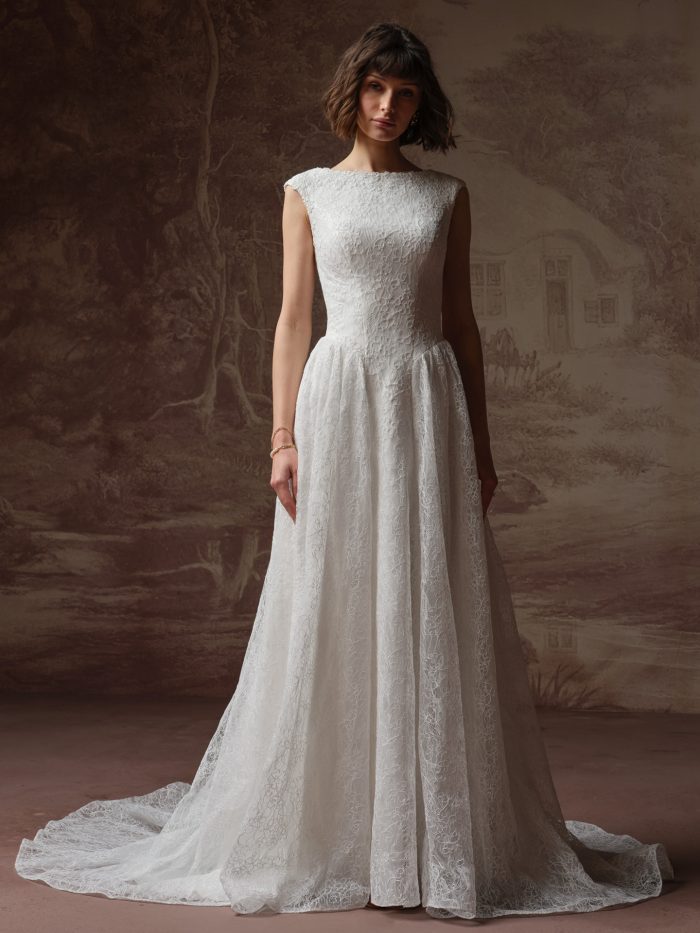 Briar A-line wedding dress by Rebecca Ingram featuring a classic bateau neckline and fitted bodice with delicate allover lace. The gown flows into a soft A-line skirt with gentle volume and a sweeping lace train, photographed against a painterly, vintage-inspired backdrop.