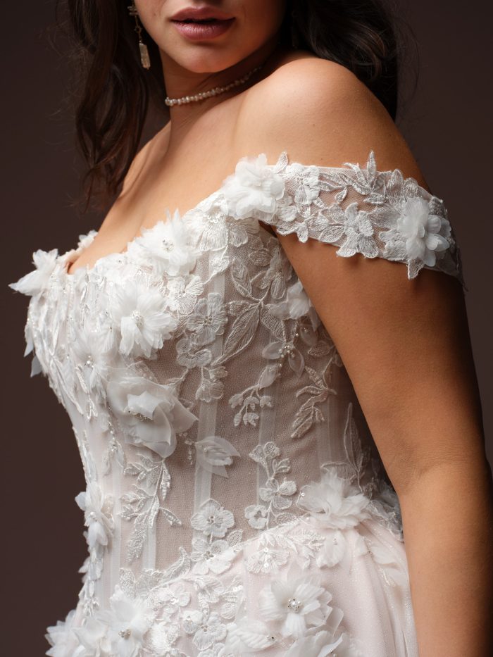 Close-up detail of the Rebecca Ingram Fleur ballgown wedding dress in ivory and blush, featuring an off-the-shoulder neckline adorned with 3D chiffon floral appliqués, embroidered lace with leaf and flower motifs, scattered pearl beading, and a sheer boned corset bodice. Styled with a delicate pearl necklace and drop earrings.