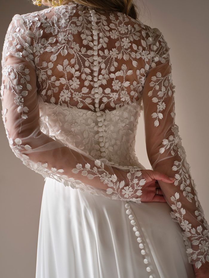 Georgina jacket accessory by Rebecca Ingram in a close-up back view, featuring sheer illusion tulle with delicate floral appliqués and a row of covered buttons running down the spine. The long sleeves are detailed with matching floral lace, layered over a fitted bodice.