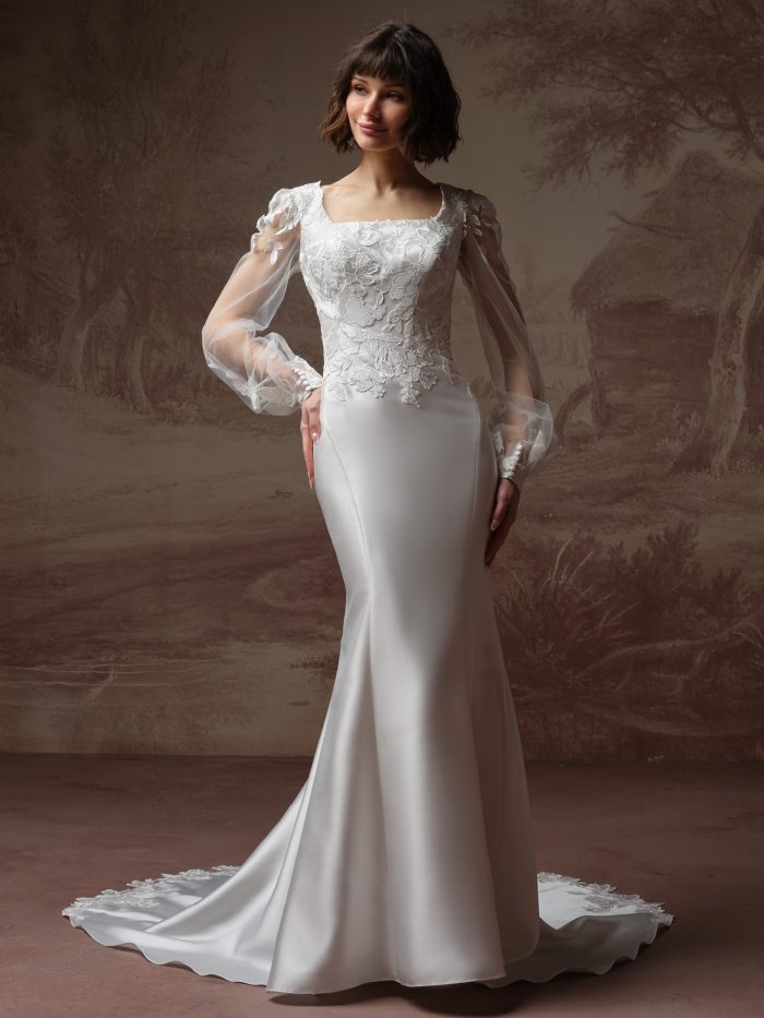 Goldie Leigh fit-and-flare wedding dress by Rebecca Ingram featuring a square neckline with floral lace appliqués across the bodice and sheer long sleeves. The fitted silhouette flares softly at the skirt, creating a clean, elegant shape against a softly painted backdrop.