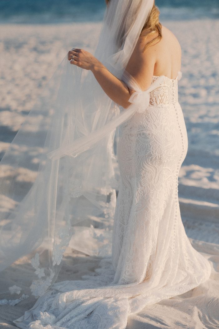 A close-up back view of a bride wearing the Berkley gown by Rebecca Ingram on a sandy beach at golden hour. The fitted lace mermaid gown features intricate geometric and floral lace patterning, off-the-shoulder tulle sleeves with a delicate bow detail, and a sweeping lace train. She holds a flowing lace-trimmed veil as ocean waves wash the shore behind her.