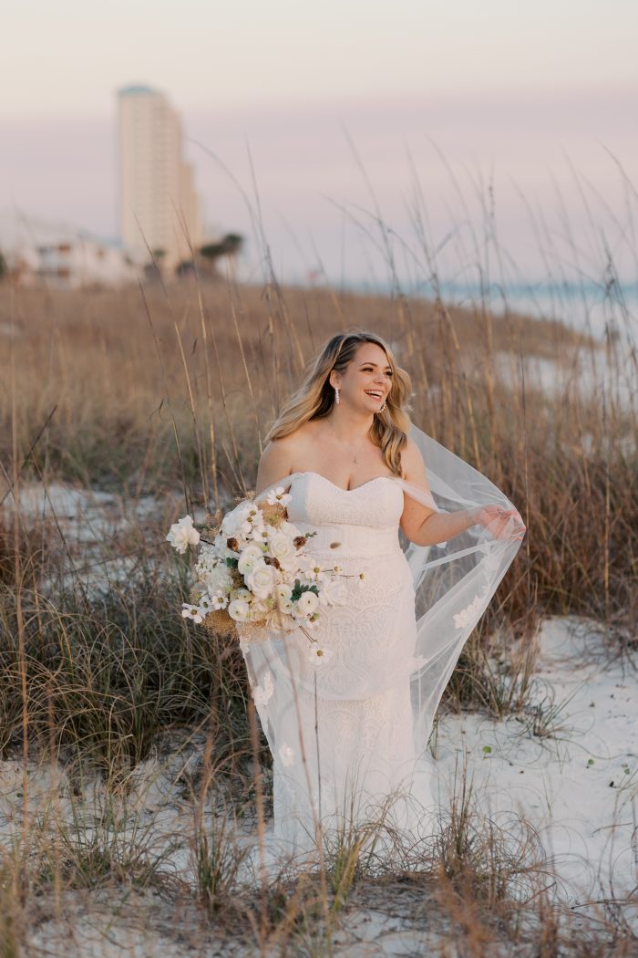 A bride wearing the Berkley gown by Rebecca Ingram laughs joyfully among sea oats on a white sand beach dune at sunset, a pastel pink and lavender sky behind her. The strapless sweetheart lace fit-and-flare gown is paired with a flowing floral-appliqué veil that catches the breeze. She carries an oversized bouquet of white roses, ranunculus, and dried botanicals, and accessorizes with drop earrings and a delicate necklace.