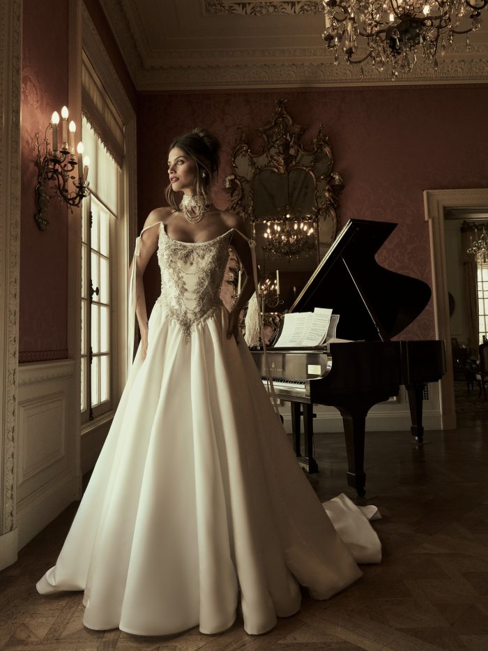 Sottero and Midgley Aelarion A-line wedding dress in ivory, featuring a square neckline with ribbon shoulder ties, heavily beaded and embroidered gold bodice, and a flowing smooth satin skirt with a chapel train. Styled with a layered pearl and crystal choker necklace. Photographed in an ornate room with a grand piano, crystal chandelier, and gilded mirror.