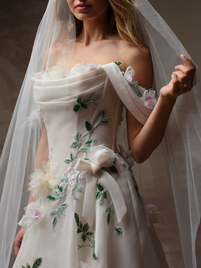 Close-up detail of the Sottero and Midgley Amaryllis wedding dress bodice, showcasing the draped organza neckline, intricate green beaded leaf embroidery, lavender floral beading, and dimensional 3D flowers in cream and blush tones with a satin bow accent at the waist. Styled with a plain cathedral veil and gold filigree earrings.