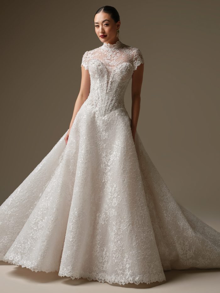 Angelica A-line wedding dress by Sottero and Midgley featuring an illusion high neckline with cap sleeves and intricate lace appliqués over a structured corset bodice. The gown flows into a full A-line skirt with shimmering lace and subtle sparkle throughout.