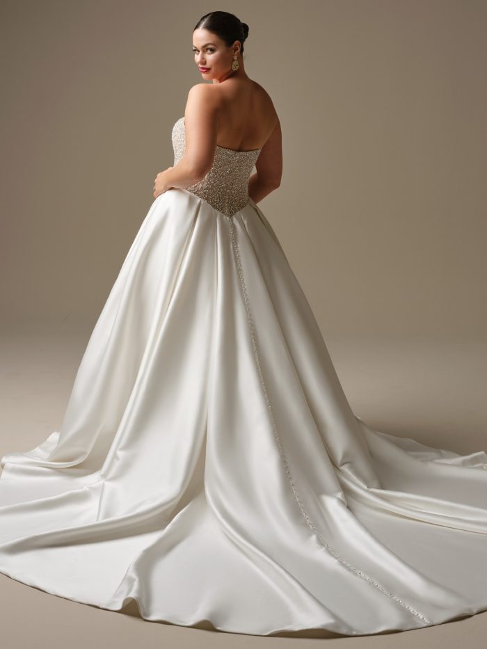 Back view of the Sottero and Midgley Dmara ballgown wedding dress in ivory on a curve model, revealing the open back with beaded corset detailing extending down the spine, and a dramatic sweeping satin cathedral train with a delicate beaded seam detail. Styled with gold drop earrings