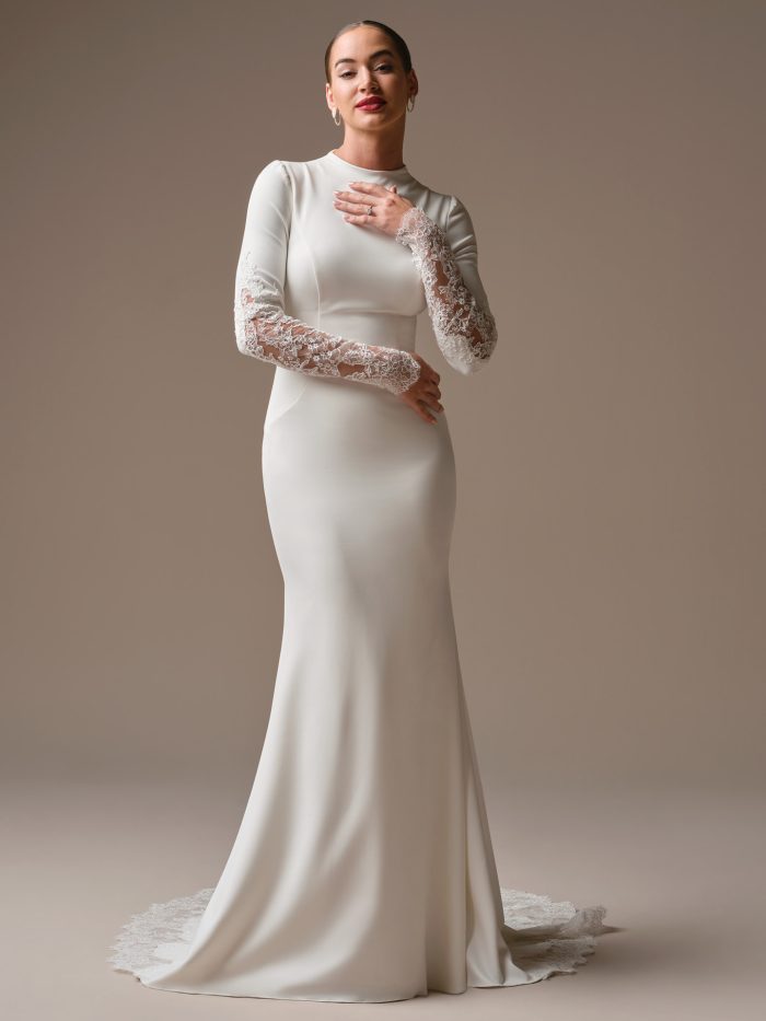 Lovai sheath wedding dress by Sottero and Midgley featuring a high neckline and sleek fitted silhouette with sheer lace long sleeves. The minimalist gown is accented with lace cuffs and a smooth skirt that flows into a soft train, styled against a neutral studio backdrop.