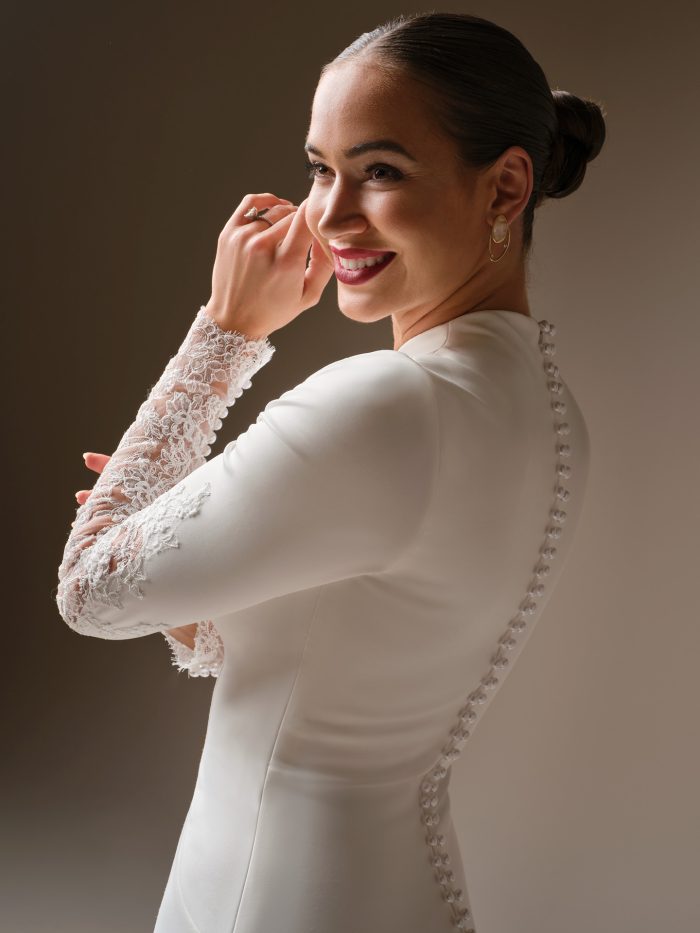 Lovai sheath wedding dress by Sottero and Midgley in a close-up view highlighting the lace sleeve detailing, covered button back, and clean, structured bodice with a high neckline.