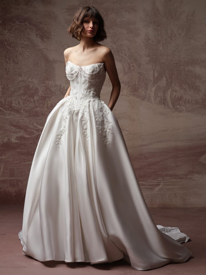 Petra ballgown wedding dress by Sottero and Midgley with a strapless, pearl-draped corset bodice and floral appliqués flowing into a full satin skirt and chapel train, shown in a full-length view against a softly textured neutral backdrop.