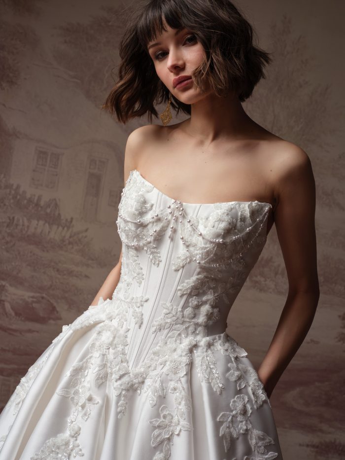 Petra ballgown wedding dress by Sottero and Midgley featuring a strapless corset bodice with pearl draping and dimensional floral appliqués, shown in a close-up view highlighting intricate beading and structured boning against a softly textured neutral backdrop.
