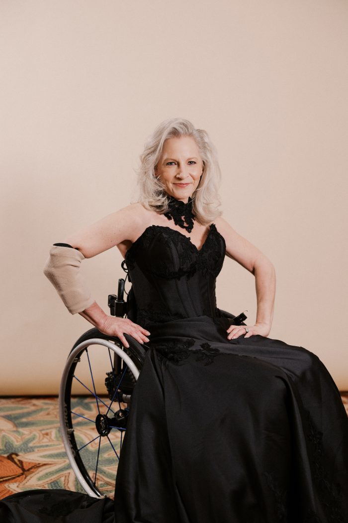 Image 2 (Tammy_0021.JPG):
A bride wearing the Serafina Lane gown by Maggie Sottero poses confidently in a manual wheelchair against a soft neutral background. The dramatic black strapless sweetheart gown features a corset-style lace bodice, a lace choker collar, scattered floral lace appliqués on the full satin skirt, and a sweeping train. She has voluminous silver-white hair, wears a prosthetic arm, and smiles directly at the camera with hands resting on the wheelchair wheels.