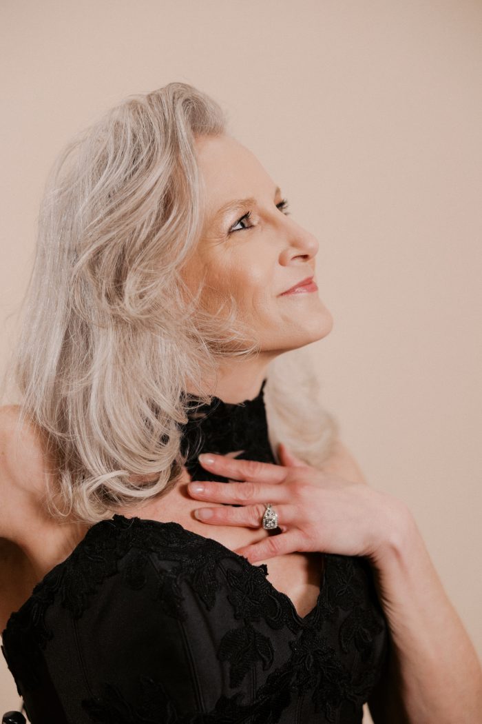 A close-up profile portrait of a bride wearing the Serafina Lane gown by Maggie Sottero, gazing upward with a serene expression. The black strapless sweetheart gown features ornate lace appliqué detailing along the bodice and a delicate lace choker neckline. She has elegant silver-white wavy hair and wears a large pear-shaped diamond ring.