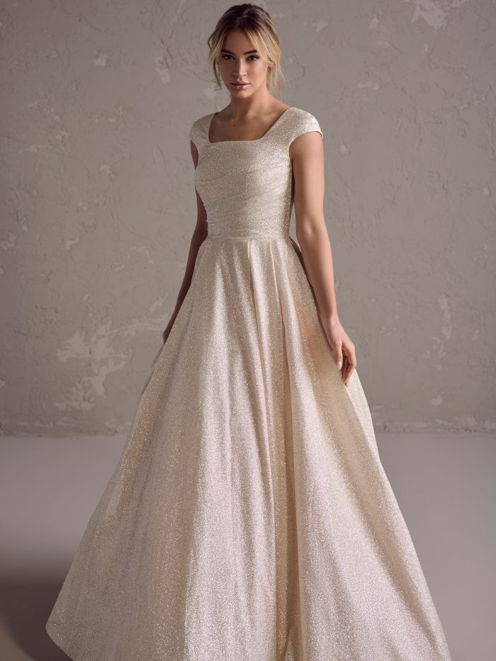 Front view of the Anniston Leigh ball gown wedding dress by Maggie Sottero, featuring a modest square neckline with cap sleeves, ruched bodice, and a full shimmer satin ball gown skirt with train.