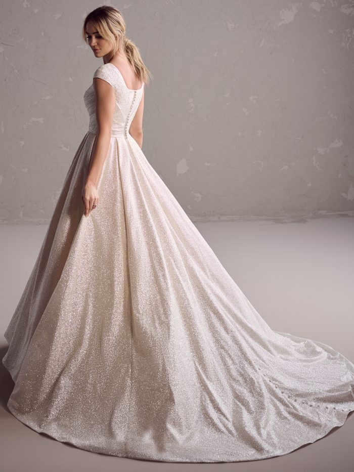 Back view of the Anniston Leigh ball gown wedding dress by Maggie Sottero, showing a modest square back with cap sleeves and covered button closure down the full shimmer satin ball gown skirt and train.