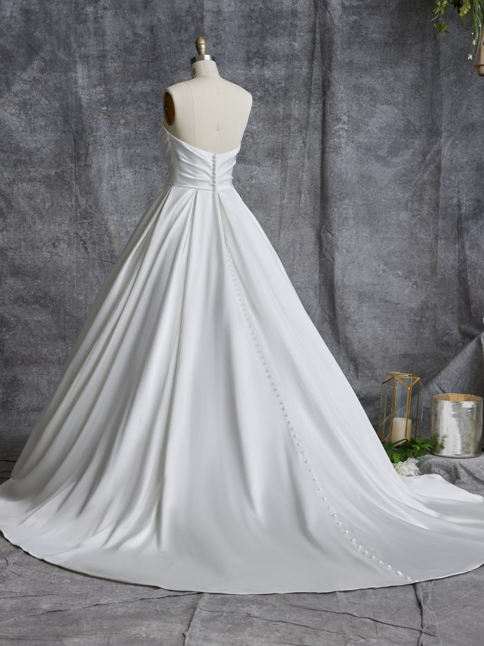 Back view of the Anniston Marie ball gown wedding dress by Maggie Sottero on a dress form, showing a strapless sweetheart back with covered button closure down the full satin ball gown skirt and train.