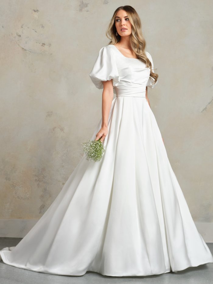Front view of the Anniston Marie Leigh ball gown wedding dress by Maggie Sottero, featuring a modest square neckline with puffed short sleeves, ruched bodice, and a full satin ball gown skirt with train, styled with a small bouquet.