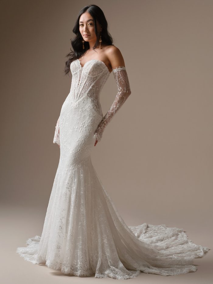 Front view of the Bellevue fit-and-flare wedding dress by Maggie Sottero, featuring a deep plunging lace bodice with off-the-shoulder straps, detachable lace opera gloves, and a fitted mermaid lace skirt with train.