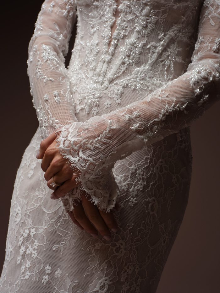 Close-up of the Bellevue Royale gown by Maggie Sottero, showing the plunging lace bodice and beaded lace sleeve with hands clasped.
