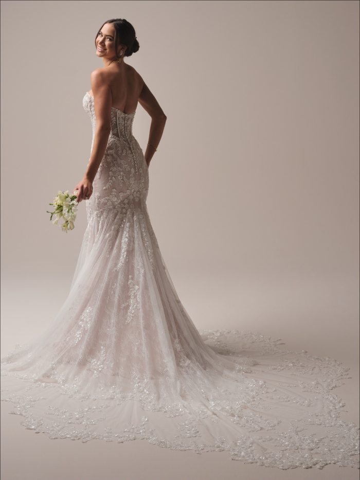 Maggie Sottero Benjie Royale Fit and Flare wedding dress in ivory, back view showing open low back with sheer lace corset detail, body-skimming beaded lace silhouette flaring at the hem, and sweeping cathedral train with scalloped lace border.