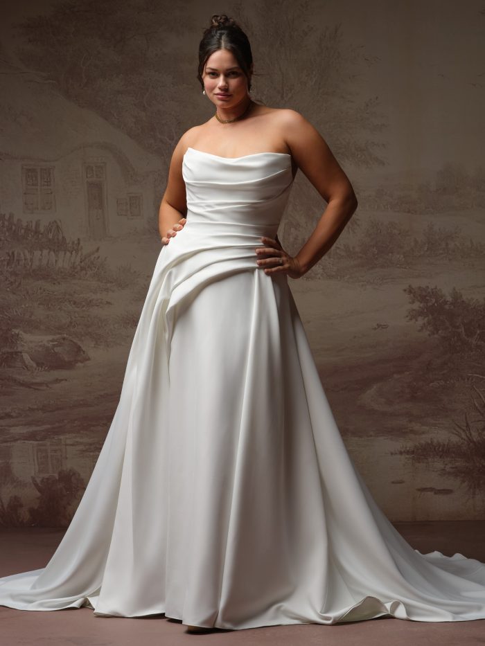 Maggie Sottero Calliope A-line wedding dress in ivory, curve fit — strapless sweetheart neckline with draped bodice, worn by a plus-size model with hands on hips, full satin skirt with sweeping train.