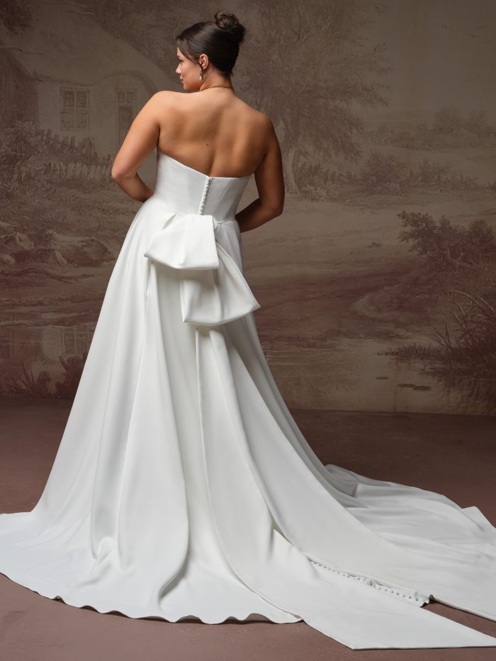 Maggie Sottero Calliope A-line wedding dress in ivory, curve fit — back view showing strapless bodice with covered button closure, oversized satin bow at the waist, and cathedral-length train.