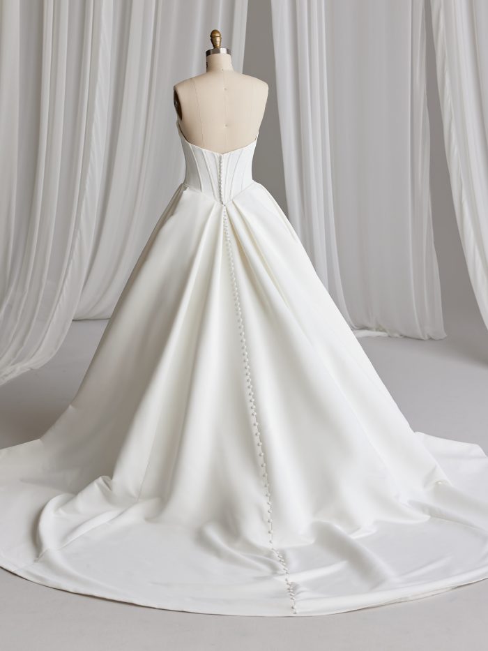 Back view of the Derrick ball gown wedding dress by Maggie Sottero on a dress form, showing a low sweetheart back with covered button closure down the full satin ball gown skirt and train.