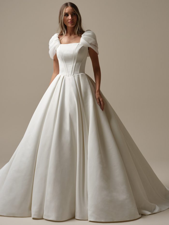 Front view of the Derrick Leigh ball gown wedding dress by Maggie Sottero, featuring a square neckline with draped off-the-shoulder puff sleeves, structured seamed bodice, and a full satin ball gown skirt with train.