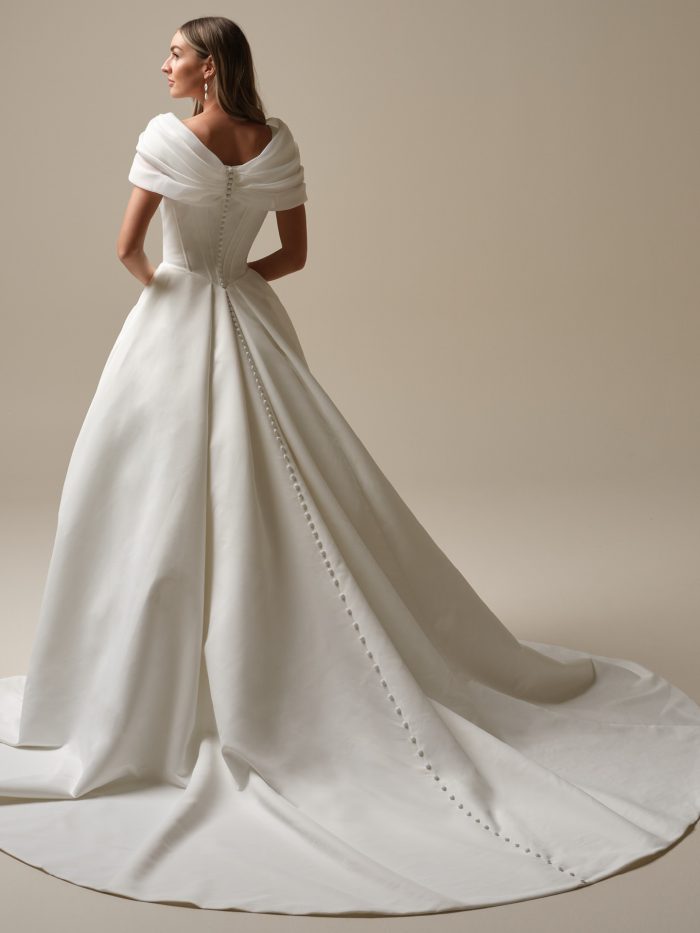 Back view of the Derrick Leigh ball gown wedding dress by Maggie Sottero, showing draped off-the-shoulder sleeves, a corset back with covered button closure, and a full satin ball gown skirt with train.