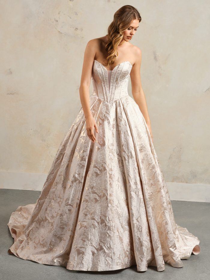 Front view of the Derrick Marie ball gown wedding dress by Maggie Sottero, featuring a deep plunging strapless neckline with structured seaming and a full metallic floral jacquard ball gown skirt with train, in a blush colorway.